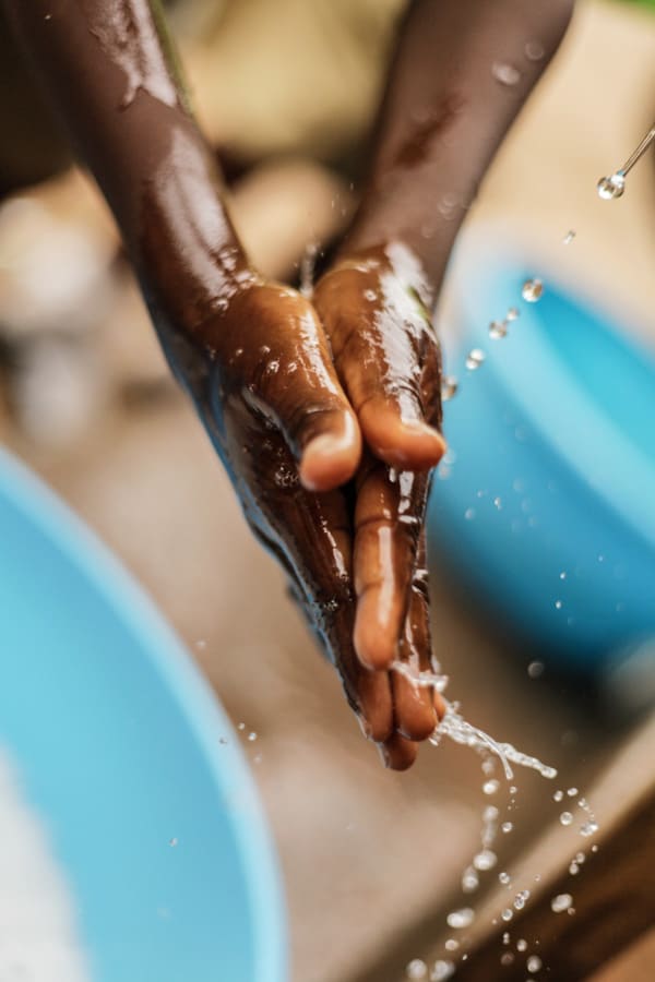 A boy is washing his hands with clean water. The suds rinse and drain into a blue tub.