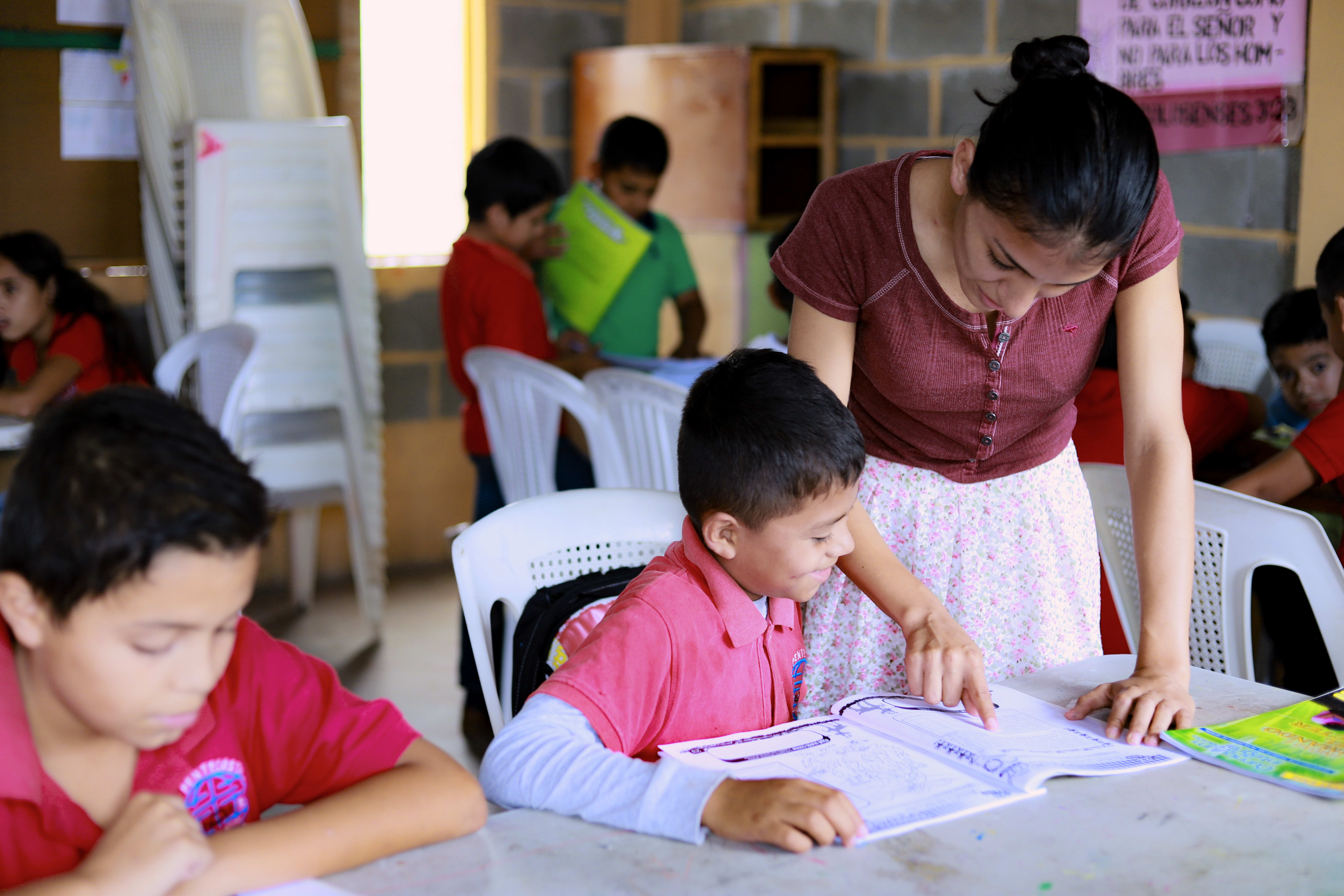 A tutor, Debora, helps a young boy, her student, with a letter he is composing in his notebook.