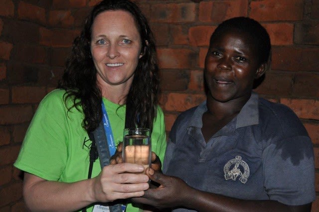 A woman in a bright green shirt stands beside a woman in a grey shirt and holds a cup of water.