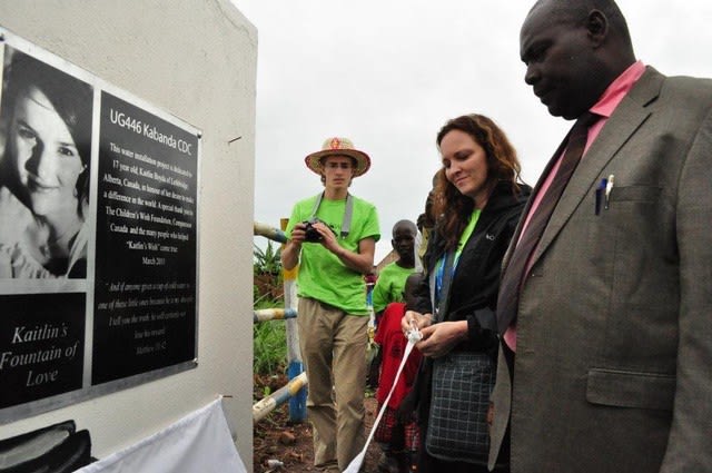 A man in a suit, a woman in a black jacket and a man in a green shirt and straw hat stand in front of a well with a dedication plaque.