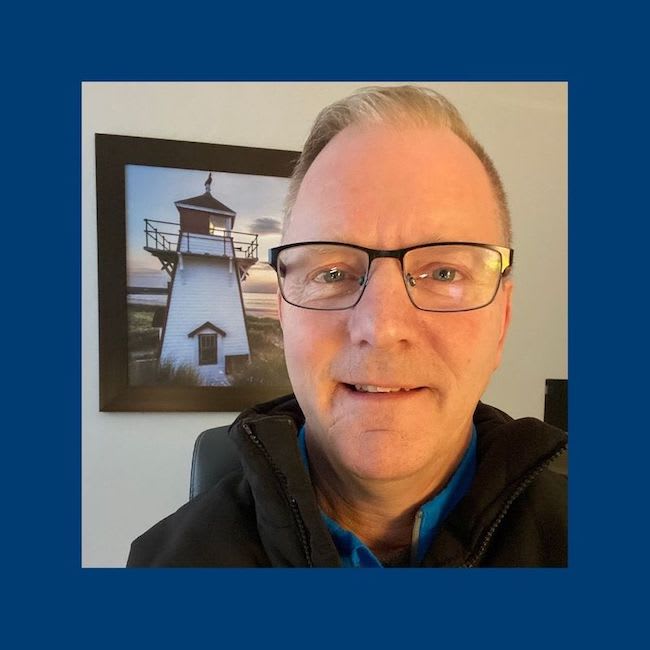 Pastor Jay wearing glasses and a dark jacket, in front of a photo of a lighthouse.