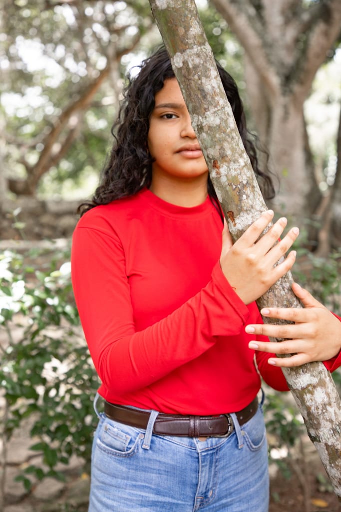 A young girl is standing behind a tree in a bright red shirt and blue jeans.