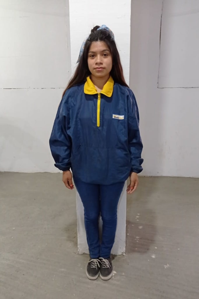 A picture of Ana, Gail's sponsored child. She is standing against a white wall smiling. She is wearing a dark blue raincoat with a yellow collar and blue jeans.