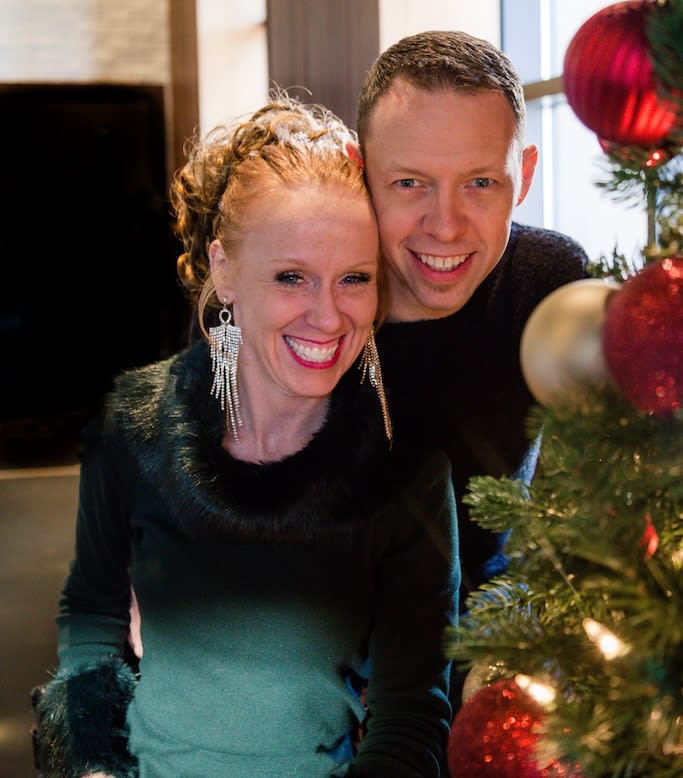 Vahen is pictured with her husband in front of a Christmas tree. They are both smiling. Vahen is wearing a dark green sweater.