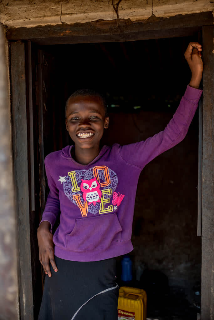 A girl wearing a purple shirt, stands in a doorway and smiles.