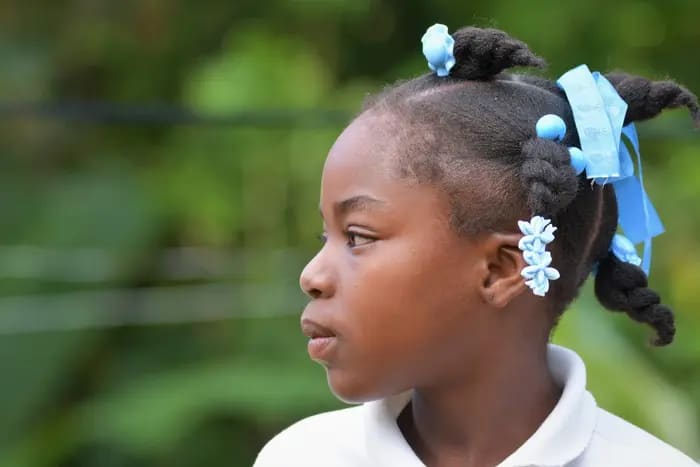 A girl looks to the side. She has blue bows in her braided hair.