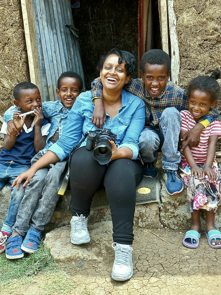 Woman holds a camera and sits on the front steps of a home. There are four kids around her laughing.