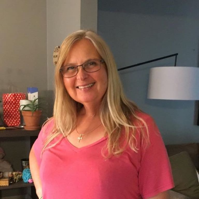 A woman stands in a pink t-shirt smiling. She has blonde hair and glasses.