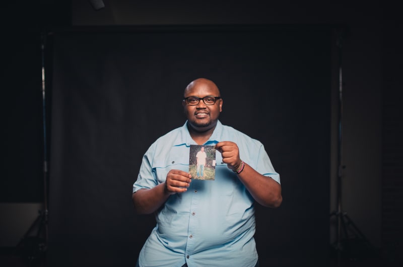Owen, an alumni from Kenya, is wearing a light blue shirt. He is holding up a picture of himself as a child