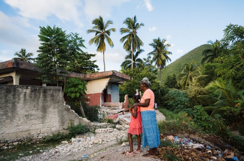 A girl in pink and a grandmother in pink and blue stand in front of a damaged home and rubble