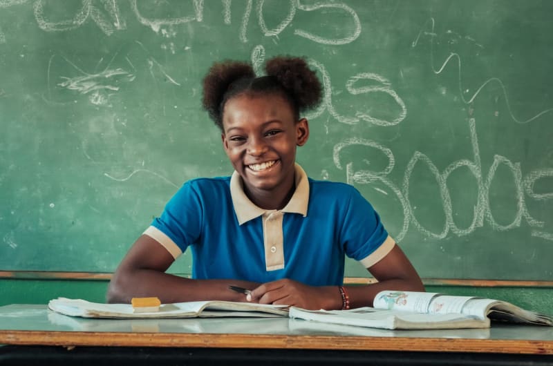 A young girl sits at a desk with books in front of her and smiles