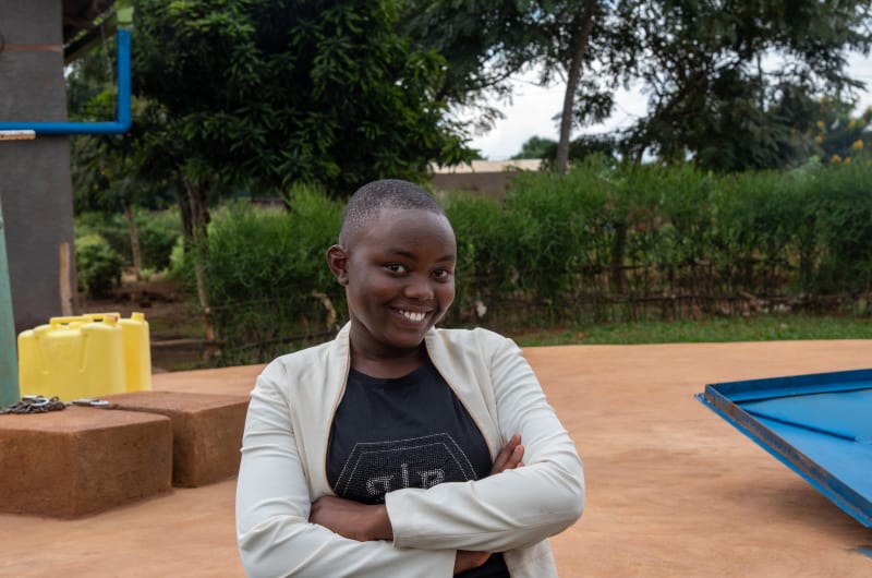 Emeline is wearing a black dress with a white sweater. She is standing with her arms crossed in front of her. There are trees and bushes behind her.