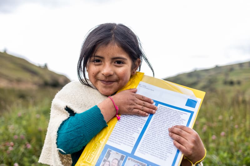 A young girl with brown hair stands outside while holding a letter in an embrace.