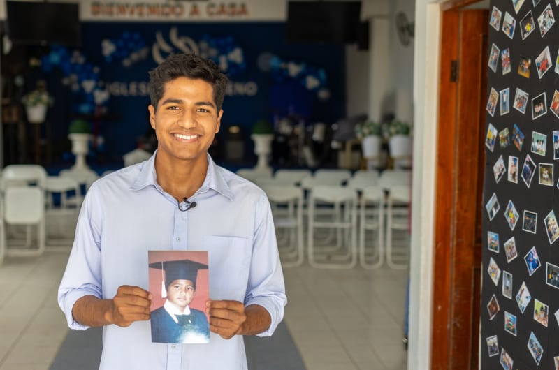 A young man stands in a blue button-up shirt holding a picture of himself as a young boy.
