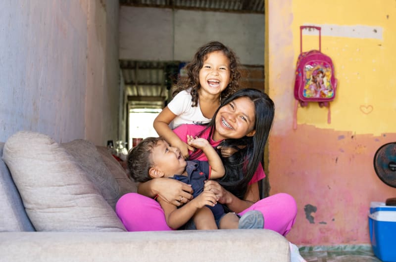 In Brazil, Gabriella and her children, Dávilla and Ithallo, are smiling and laughing as they play together on the sofa.