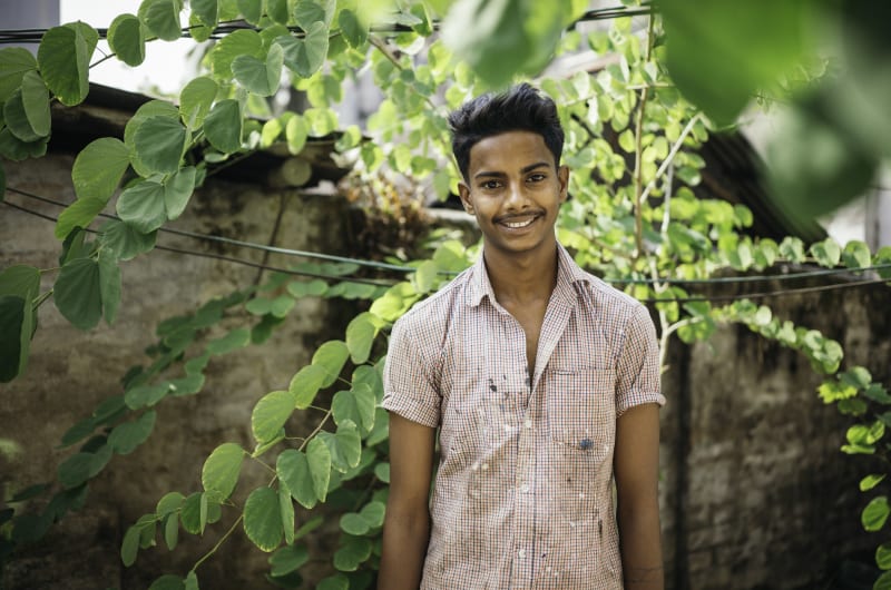 Shanto stands amidst green leaves in a plaid shirt and smiles.