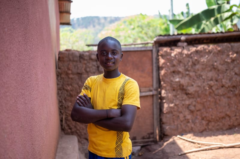 Patrick, wearing yellow, poses in front of his house with his arms crossed and a smile.