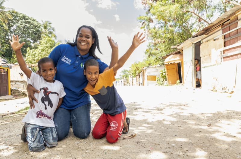 Sugeli, an administrative assistant at a Dominican Republic Compassion centre, poses with two boys on the street. They are kneeling and smiling while throwing their hands up.