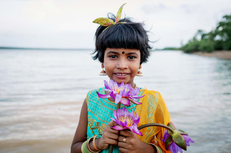 A young girl stands on the shore of the ocean in a colourful dress. She's holding a pink flower.
