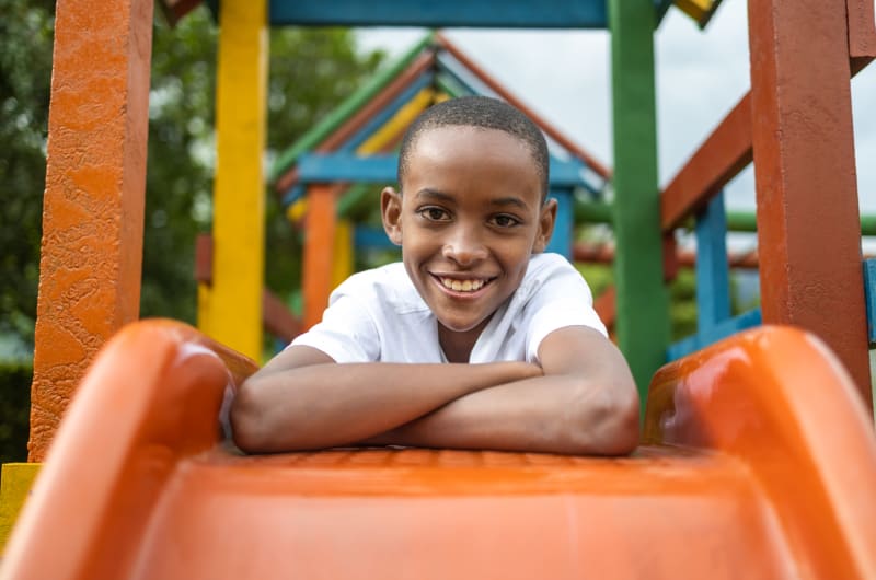 A young boy in a white tshirt leans over an orange slide with his arms crossed. He is smiling.