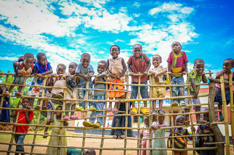 A group of smiling children gather around and climb up a fence.
