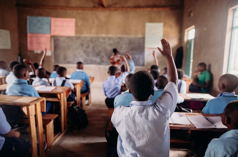 Students in a classroom raise their hands to answer a question the teacher has written on the board.