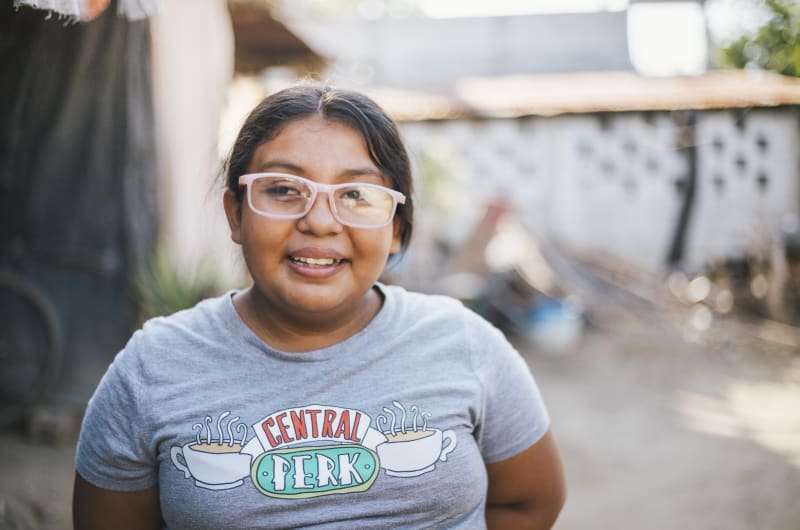 A girl wearing pink glasses and a grey t-shirt smiles.