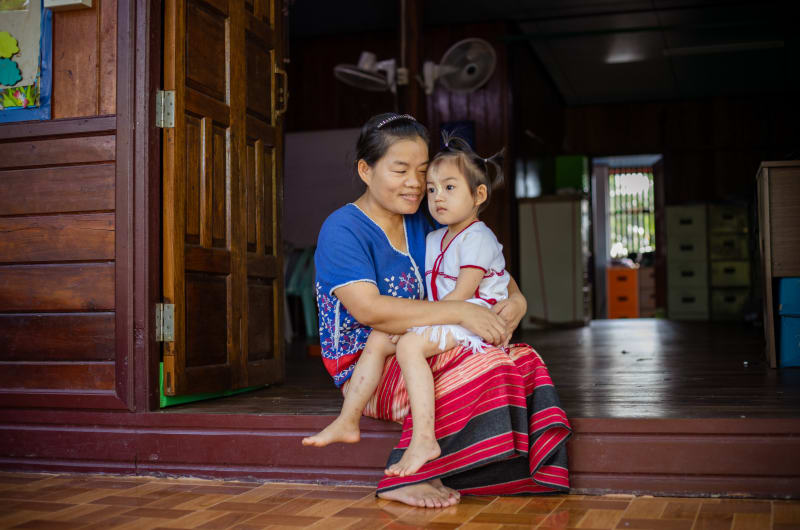 A mother in a blue and red dress sits on the front stoop holding her daughter on her lap. The daughter wears white.