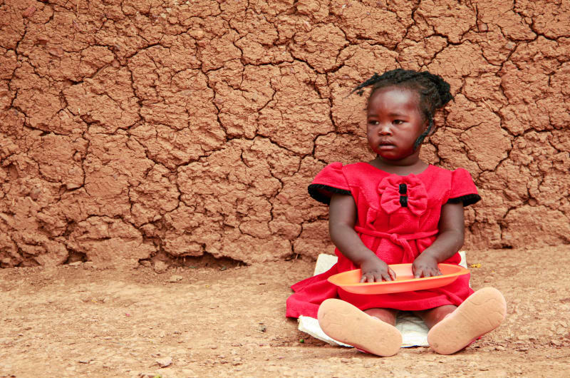 A little girl in a red dress sits against a mud wall and holds an empty plate.