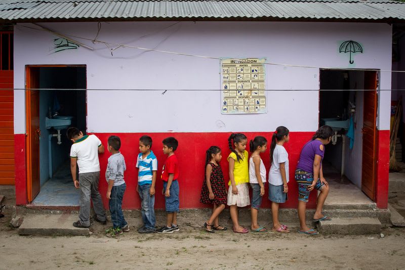 A group of children wait in single file lines outside the white and red painted washrooms.