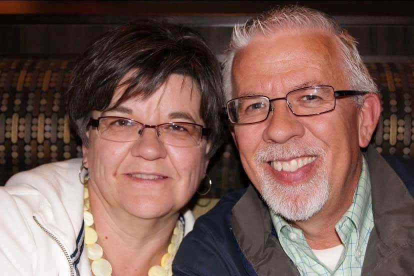 A woman with glasses and a yellow necklace and a man with glasses smile together.
