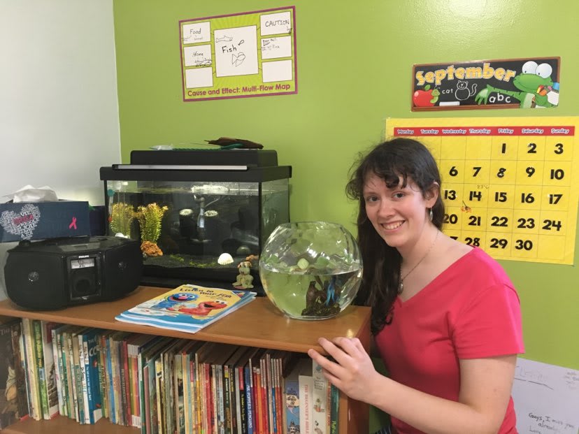 A woman in a red tshirt stands in a green classroom beside a fishbowl