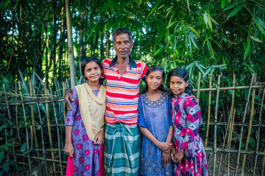 Bobita standing with her father, Manno, and her two younger sisters, Kobita (left) and Sobita (right). They are standing in front of a forest.