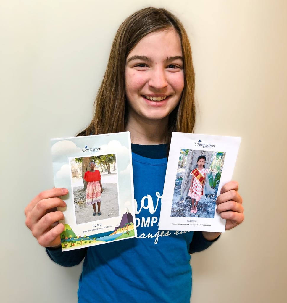 A girl in a blue shirt holds up to pictures and smiles at the camera